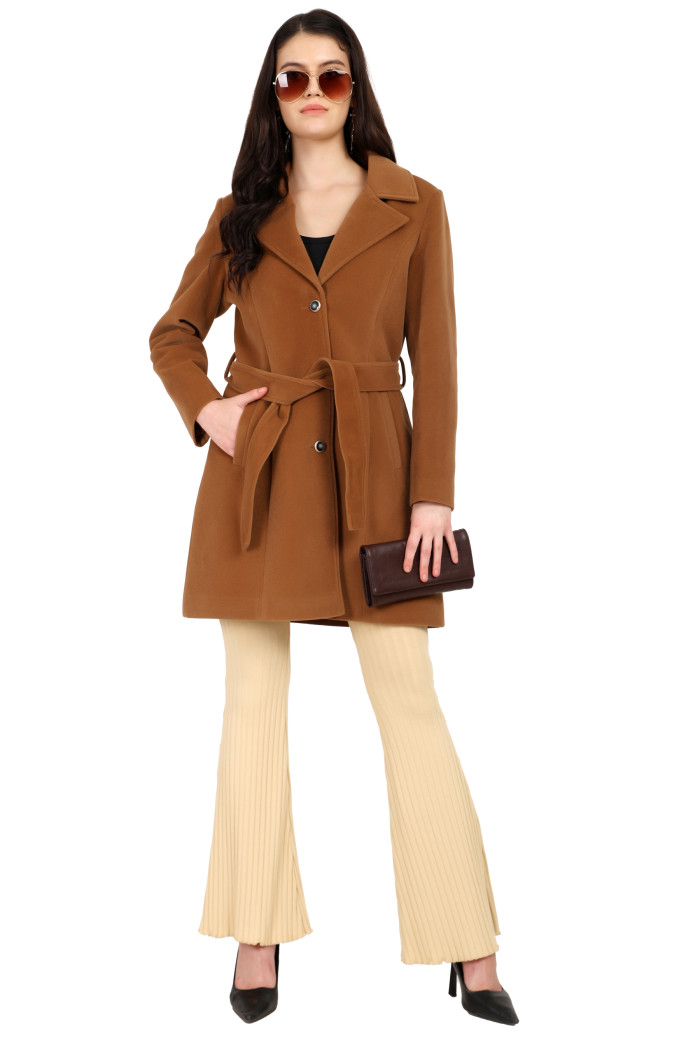 A standing pose of a woman in black shades, wearing Trufit’s Long Dk. Camel Velvet coat with a lapel collar, button closure, belt, side pockets, and skin bell bottom, holding a clutch from her left hand and her right hand in the pocket.